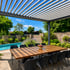 Große Holztafel mit acht schwarzen Stühlen unter einer Pergola neben einem Pool und Garten.