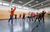 Ein Mann im orangefarbenen Shirt leitet eine Gruppe von Kindern beim Sport in einer Turnhalle an.