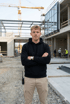 Junger Mann mit dunklem Kapuzenpullover und beiger Hose steht mit verschränkten Armen auf einer Baustelle.