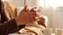 A person with dark skin clasps their hands together while sitting on a couch near two coffee mugs.