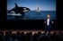 Man in a suit speaks on stage to an audience, with a screen behind him showing an orca and a sailboat.