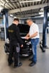 Zwei Kfz-Mechaniker begutachten ein Dokument vor einem schwarzen Auto mit geöffneter Motorhaube in einer Werkstatt.