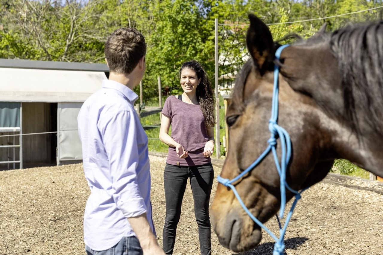 Seraina im Gespräch mit einem Klienten auf dem Reitplatz, im Vordergrund ein braunes Pferd