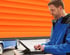 Ein Techniker in blauer Arbeitskleidung bedient einen Laptop vor einem orangenen Rolltor und einem blauen Fahrzeug.