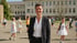 Frau mit kurzen Haaren und schwarzem Blazer lächelt vor einem Schulgebäude mit spielenden Kindern.