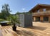 Grauer Container vor einem Holzhaus auf einer Terrasse mit Whirlpool und Palme.