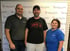 Three people stand smiling in front of a PC Medical Centers backdrop. The center man wears a black shirt reading "The Brick."