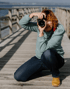 Eine Frau mit rotem Haar kniet auf einem Holzsteg und fotografiert mit einer schwarzen Kamera.