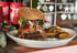 Ein großer, doppelter Burger mit Speck und Käse, daneben Kartoffelecken, auf einem weißen Teller.