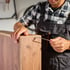 Mann in kariertem Hemd und Arbeitslatzhose montiert braunes Holzregal mit Schraubendreher und Schraube.