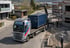 Ein LKW mit Ladekran entlädt einen blauen Container auf einer Baustelle neben einem Rohbau.