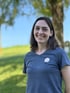 Lächelnde Frau mit schulterlangem braunem Haar und blauem T-Shirt mit kleinem weißen Logo im Freien.