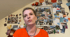 Eine Frau mit orangefarbenem Oberteil spricht; im Hintergrund eine Wand mit vielen Fotos und Postkarten.