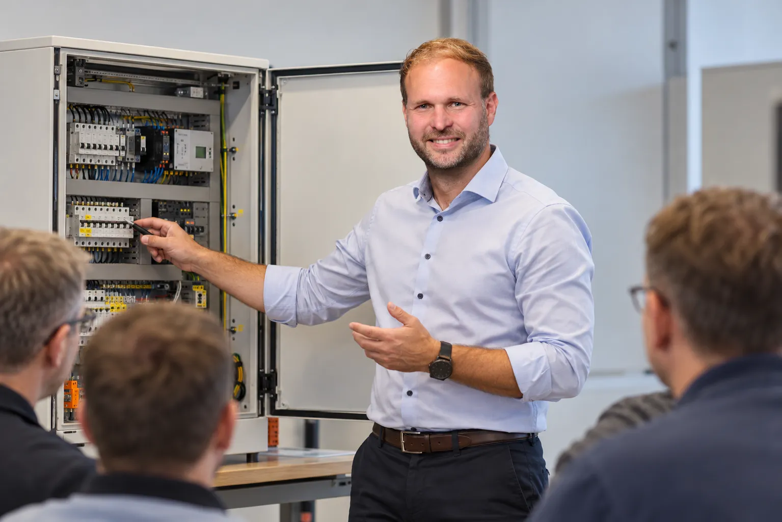 Dozent erklärt einer Gruppe von Mitarbeitern die Bauteile in einem geöffneten Schaltschrank