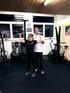 Frau stemmt Hanteln im Fitnessstudio, Trainer unterstützt sie.