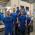 Sechs Mitarbeiter in blauen Hemden und Hosen vor einer großen weißen Hermle C 62 U Werkzeugmaschine.