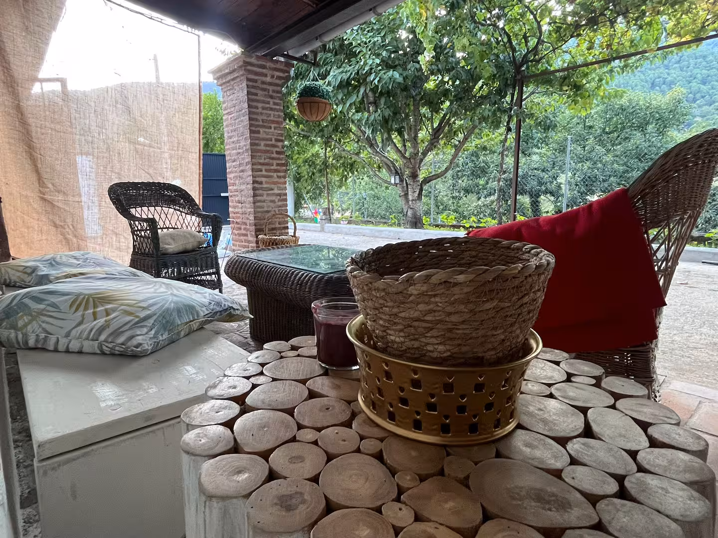 Patio exterior con muebles de mimbre, cojines estampados, una mesa de troncos cortados, y un cesto, con un fondo de árboles y montañas.