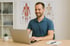 Lächelnder Physiotherapeut am Laptop mit Anatomiepostern im Hintergrund.