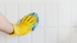 Eine Hand mit gelbem Handschuh wischt mit einem blauen Tuch eine weiße geflieste Wand ab.