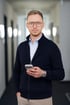 Ein Mann mit Brille und dunkelblauer Strickjacke hält ein Smartphone in einem modernen Büroflur.