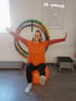 Frau macht Gymnastik auf einem orangen Pezziball mit ausgestreckten Armen vor einer Wand mit Hula-Hoop-Reifen.