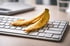 Eine gelbe Bananenschale liegt auf einer silbernen Computertastatur mit weißen Tasten auf einem Schreibtisch.