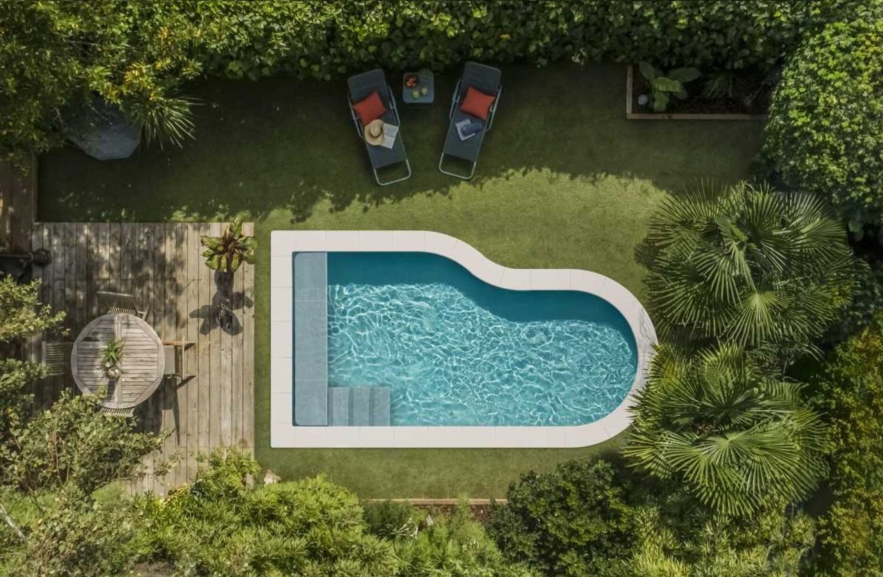 U-förmiger Pool mit Stufen, umgeben von Holzdeck, Rasen und grünen Pflanzen, von oben fotografiert.