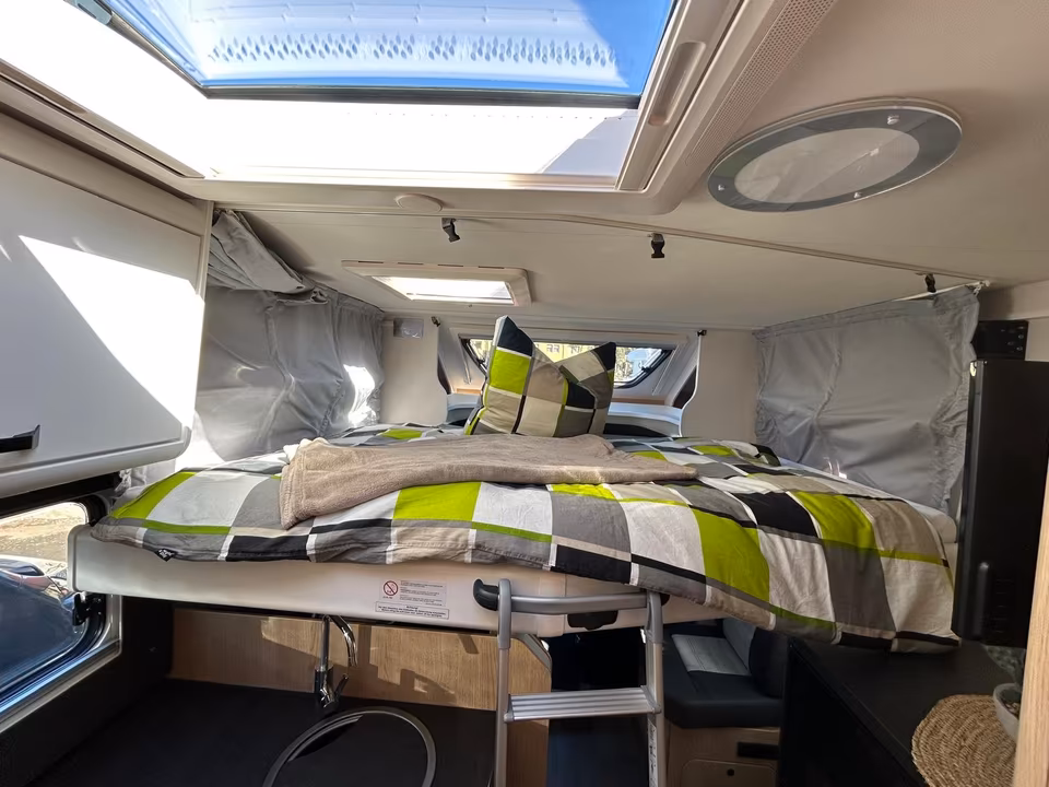Ein Schlafbereich in einem Wohnmobil mit einem hochgelegenen Bett, grauen Vorhängen und einem Dachfenster.