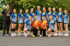 Frauen-Handballteam posiert im Freien, die meisten tragen blaue Trikots, drei vorne tragen orange Trikots.