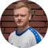 Porträt eines jungen Mannes mit kurzem roten Haar und Bart, der ein weiß-blaues Sporttrikot trägt, vor einer braunen Wand.