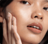 Nahaufnahme einer Frau mit glänzender Haut, braunem Lippenstift und einer Hand an der Wange für Hautpflege-Werbung.