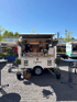 Weißer Foodtruck "Lagrein" mit zwei Personen, die Essen zubereiten, auf einem Kopfsteinpflasterplatz unter blauem Himmel.