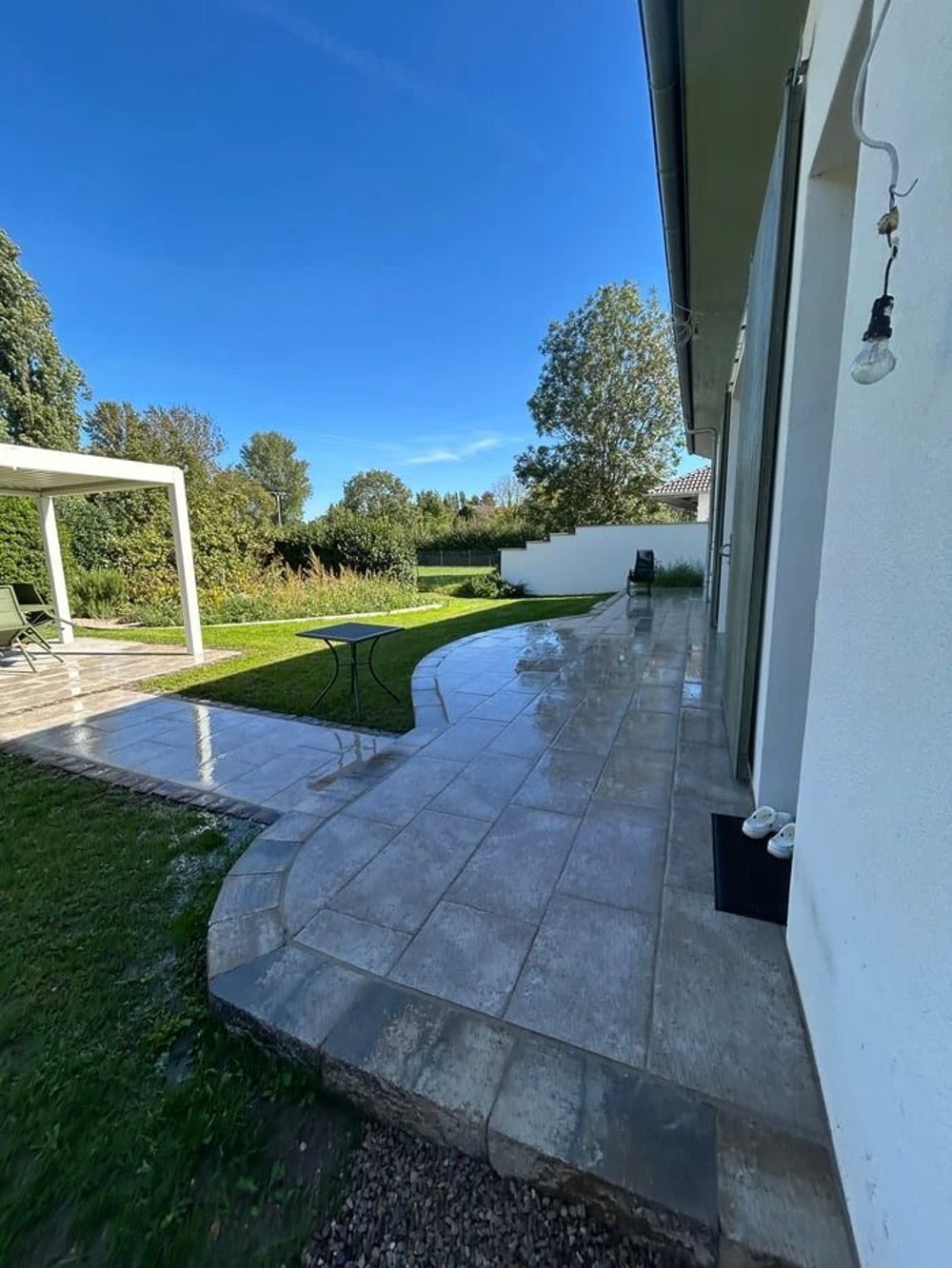 Nachher: geschwungene Terrasse aus großformatigen Platten entlang der Hauswand, Rasen und Pergola