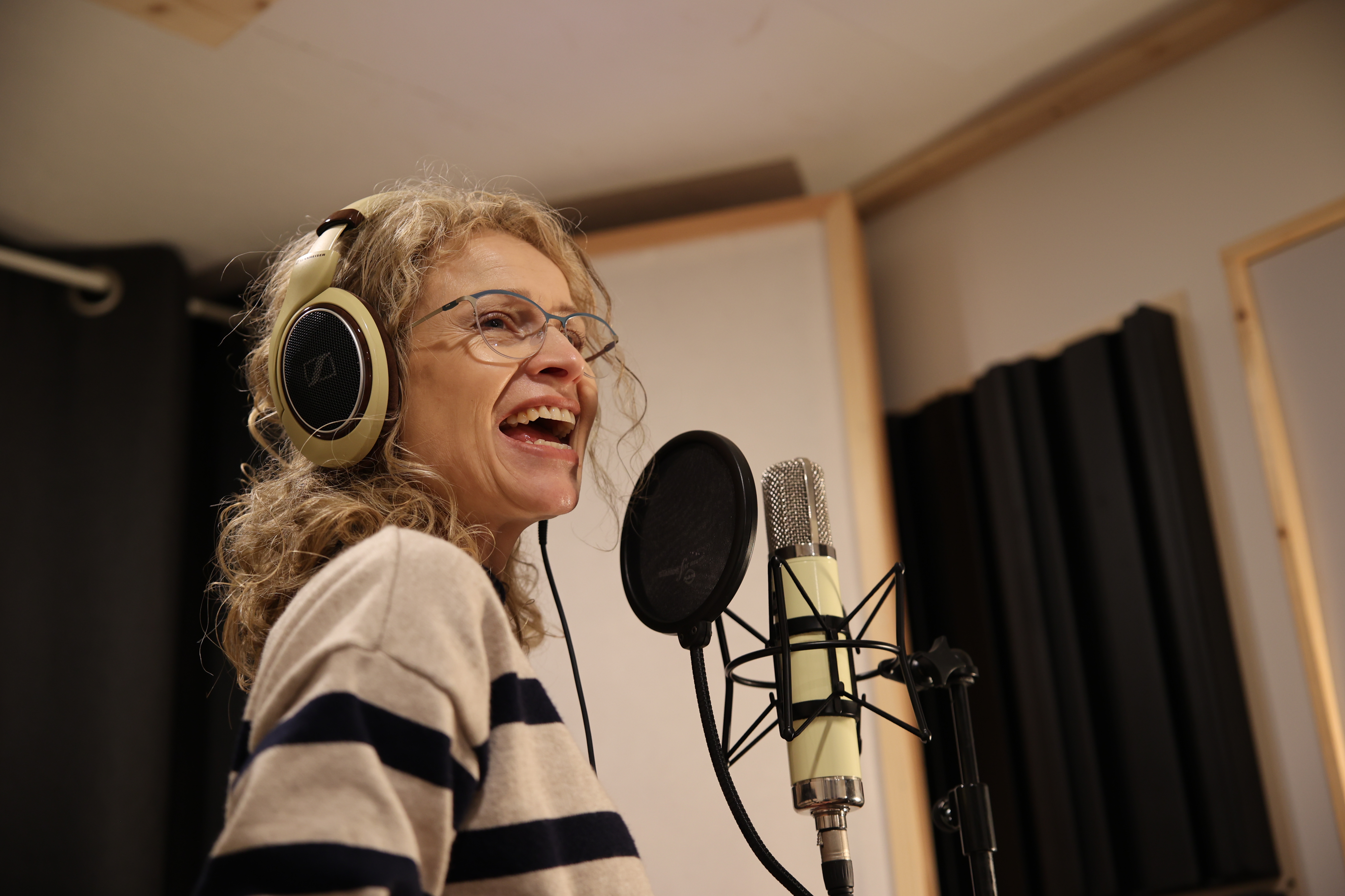 Eine Frau mit beigen Kopfhörern und Brille singt enthusiastisch in ein Mikrofon in einem Aufnahmestudio.
