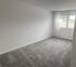 Leeres Zimmer mit weißen Wänden, grauem Teppichboden und einem Fenster mit weißen Vorhängen.