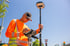 Mann in orangefarbener Warnweste und Schirmmütze bedient ein Vermessungsgerät mit GPS-Antenne unter blauem Himmel.