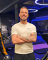 Lächelnder Mann mit Tätowierungen und T-Shirt mit Aufschrift "SANNY Personal Trainer" kreuzt die Arme.