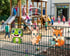 Lachender Frosch, Hase mit Karotte und Fuchs mit Gießkanne an einem Zaun vor spielenden Kindern und einem Klettergerüst.