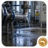 Wasser spritzt aus einem Leck in einem metallischen Industrierohr auf den Boden einer Fabrikhalle - Software für Alarm- und Gefahrenabwehrplanung - Notfall- und Krisenmanager (NKM®).