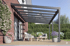 Moderne, anthrazitfarbene Terrassenüberdachung aus Aluminium über einer Holzterrasse mit Möbeln und Pflanzen.