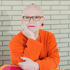 Eine Person mit Brille, orangefarbenem Pullover und roten Ohrringen lehnt an einer Ziegelwand.