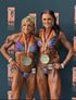 Zwei Bodybuilderinnen mit Medaillen und Startnummern 19 und 29 posieren vor einer schwarzen Wand mit NAC-Logo.