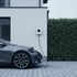 Ein graues Elektroauto lädt an einer weißen Wallbox an einer Hauswand neben einer grünen Hecke.