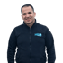 Mann in schwarzer Fleecejacke mit blauem "KIDO" Logo lächelt vor grünem Hintergrund.
