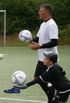 Ein Trainer hält einen Fußball, während ein Kind im Torwarttrikot einen anderen Fußball auf dem Boden berührt.