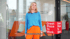 Eine Frau in blauem Oberteil und orangefarbener Hose steht vor einem Great Place To Work Schild.