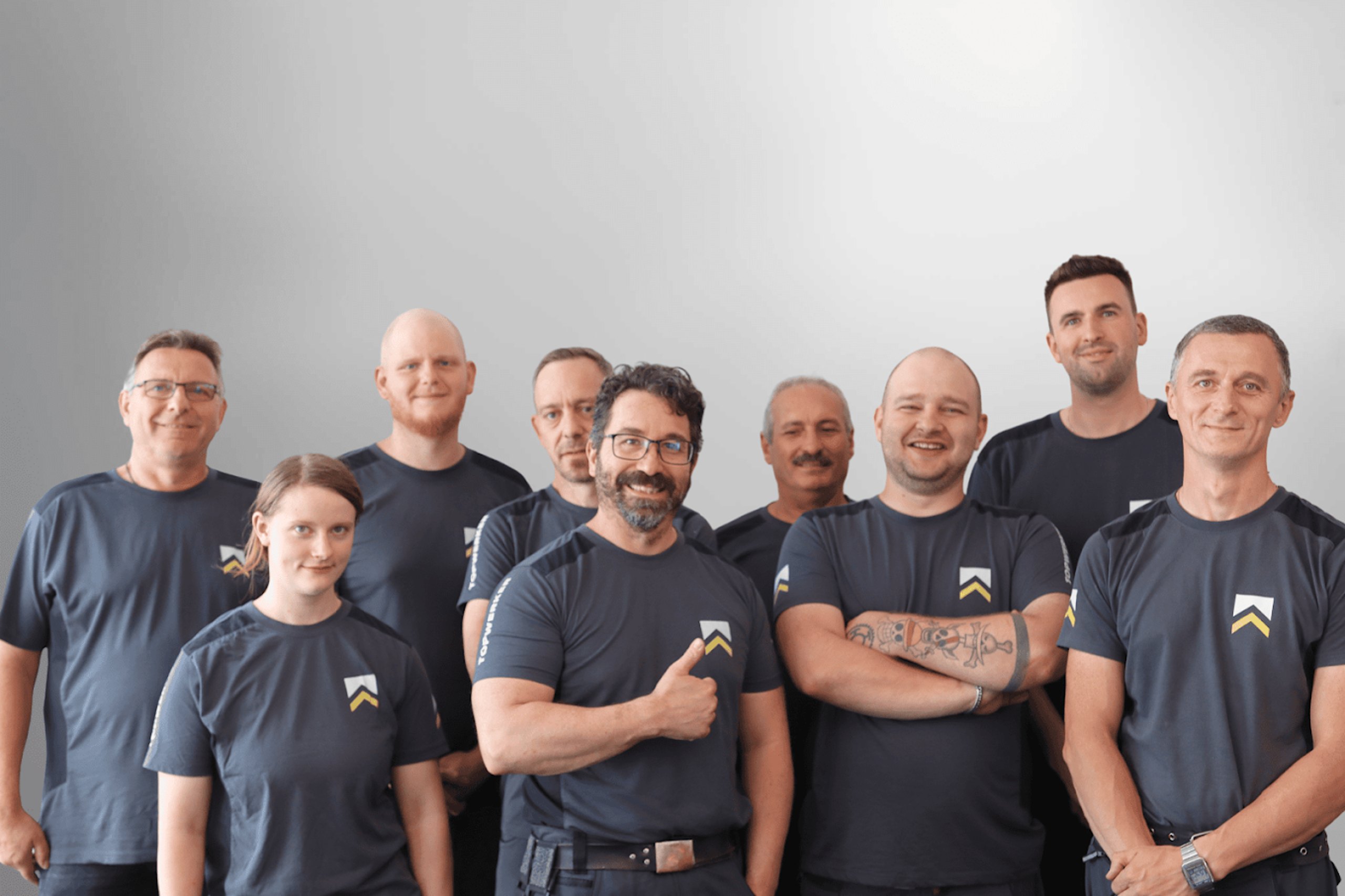 Neun Mitarbeiter in dunkelgrauen T-Shirts stehen vor grauem Hintergrund und lächeln.
