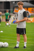 Junger Fußballspieler in graubraunem T-Shirt mit TSA-Logo und schwarzen Shorts hält blaue Markierungsteller in Händen auf dem Feld.