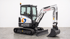 Kleiner weißer Bobcat-Bagger mit schwarzer Kabine und Schaufel auf Betonboden vor weißer Wand.