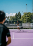 Tennisspieler auf einem lila Hartplatz steht am Netz mit Schläger und Ballkorb vor einer HEAD-Werbewand.